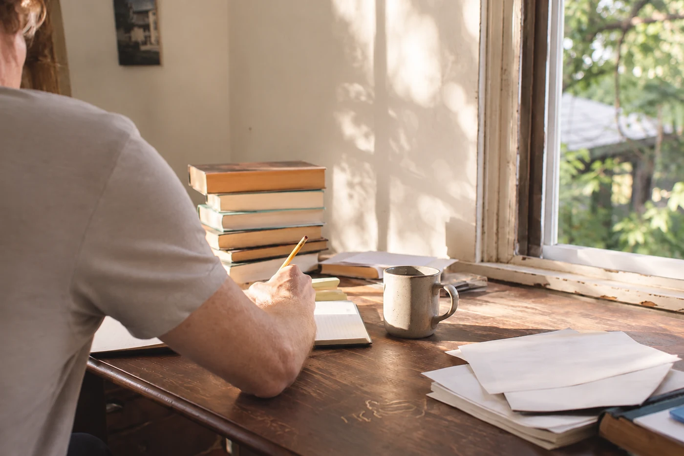 A writer works beside a sunlit window with books, loose pages, and coffee