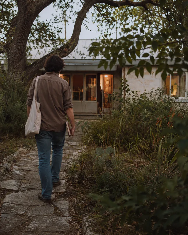A writer carrying a notebook walks toward a warmly lit Austin workshop