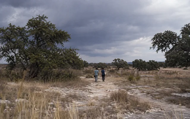 Two people walk through a rocky Texas landscape beneath storm clouds
