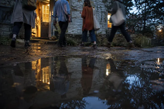 Writers leave a stone house after rain, reflected in a puddle