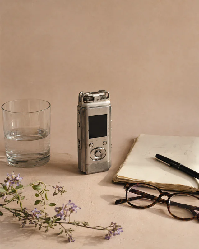 An audio recorder, notebook, glasses, water, and wildflowers arranged for a conversation