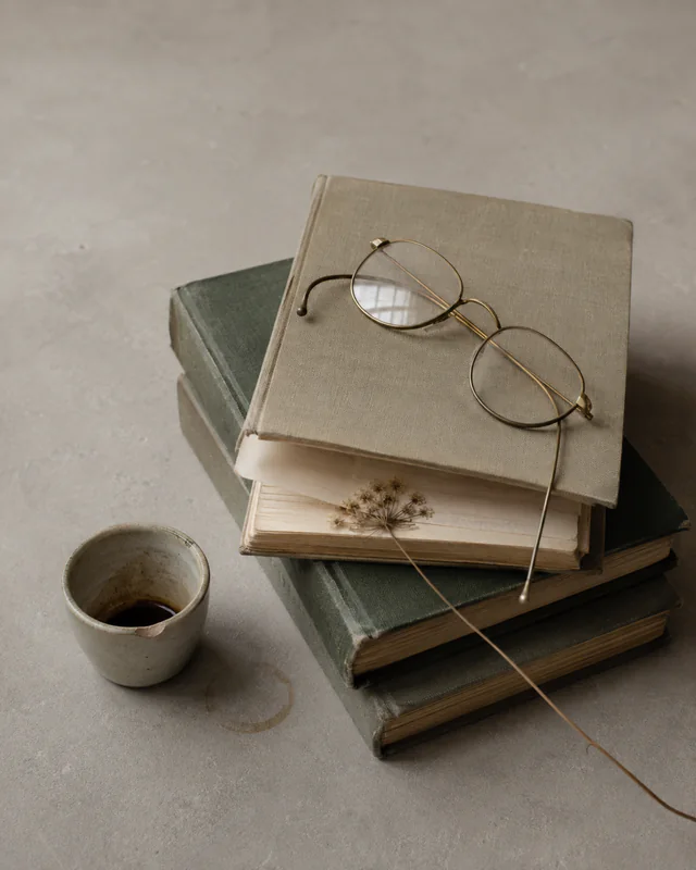 A stack of worn books with reading glasses, coffee, and a dried flower