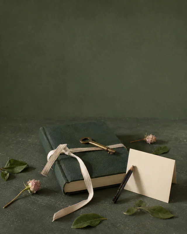 A brass key, tied notebook, note card, pencil, and flowers on a green table