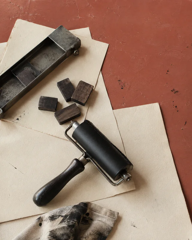 A letterpress roller, wooden type, paper, and ink cloth on a rust-colored table