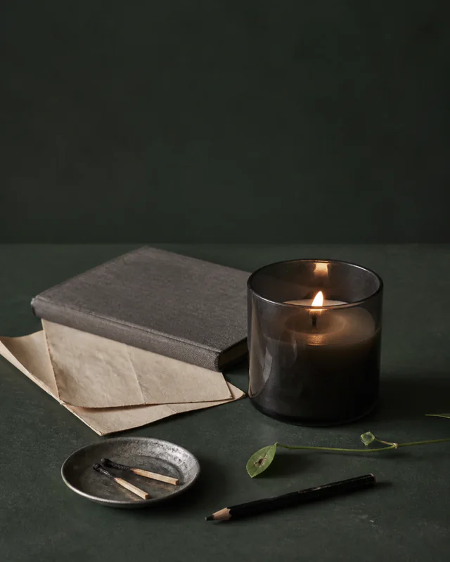 A smoked-glass candle beside a closed notebook, pencil, and small dish