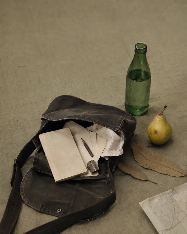 A traveling satchel packed with notebooks beside a map, pear, and green bottle