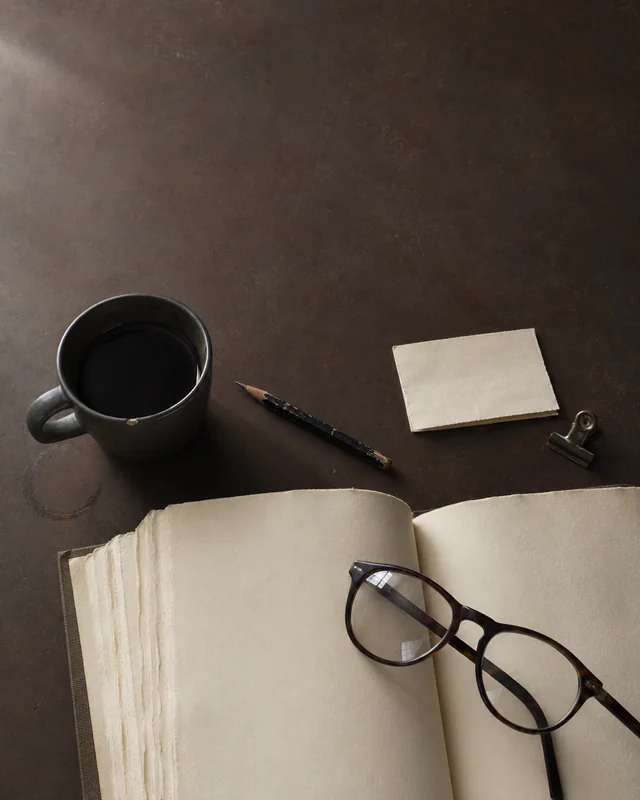 An open book with glasses, a black coffee mug, pencil, and clipped note